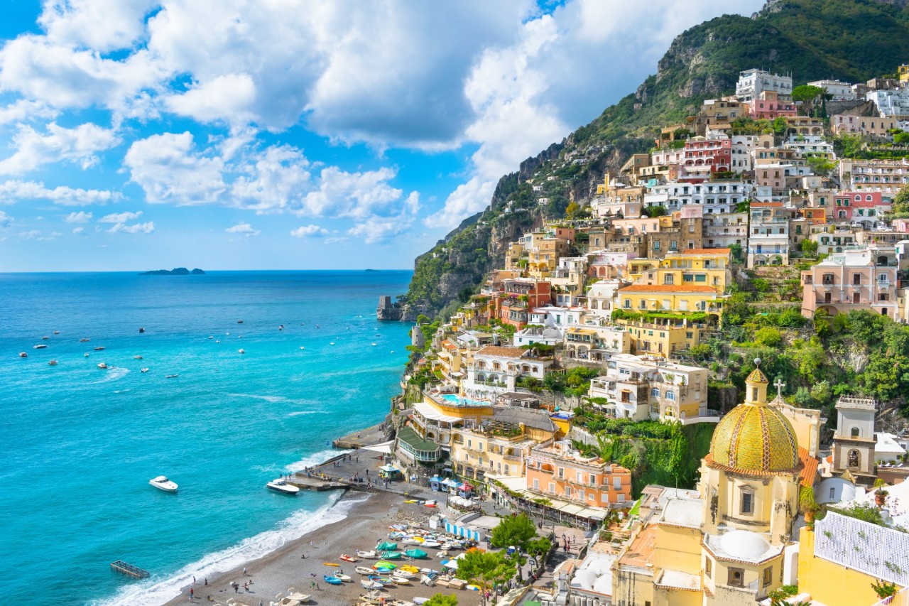 View of a town with colourful buildings that rises up on a slope directly from the turquoise sea. Below the town is a beach with numerous small boats lying on the sand.