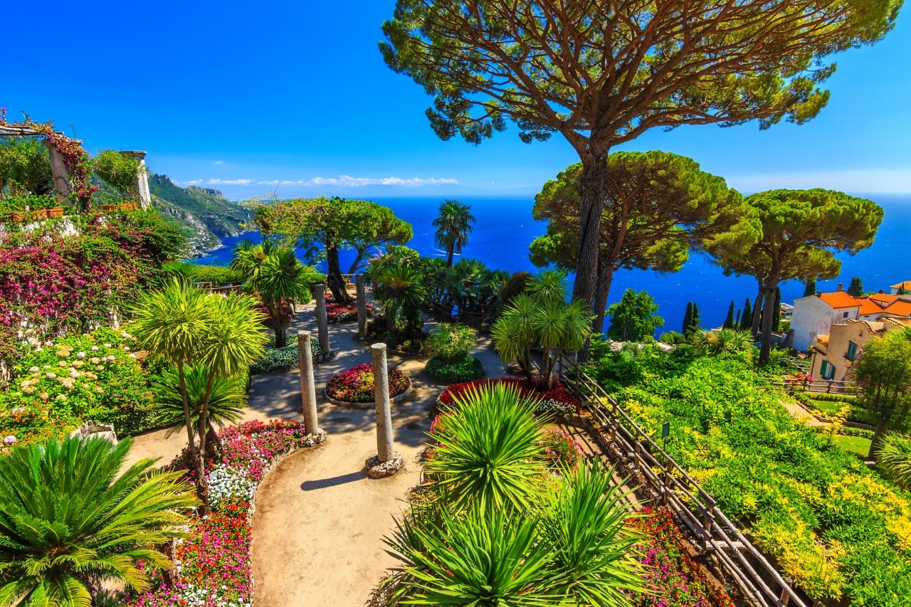 View of a landscaped garden on a hill above the sea, which is bright blue in the background. The garden consists of many different types of palm trees, flowering bushes, trees and flowers.