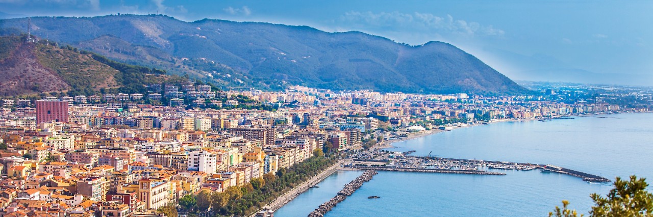 Aerial view of a large town by the sea with a spacious harbour built into the sea. A mountain range can be seen in the background.