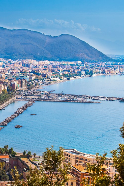 Aerial view of a large town by the sea with a spacious harbour built into the sea. A mountain range can be seen in the background.