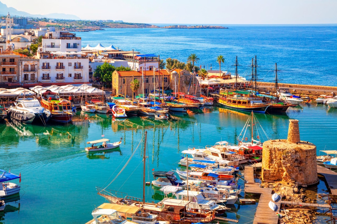 Aerial view of a small harbour, enclosed to the open sea, in which numerous small, colourful boats are moored. A small boat leaves the harbour. Multi-storey houses rise behind the harbourfront. A long stretch of coastline can be seen behind the houses.