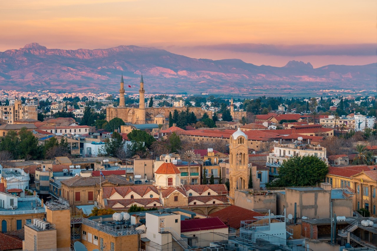 Aerial view of a city in the evening light. Numerous historic buildings with tiled roofs, houses and several churches as well as a mosque with two minarets are visible in the background. A mountain range can be seen in the background, glowing red in the evening light.