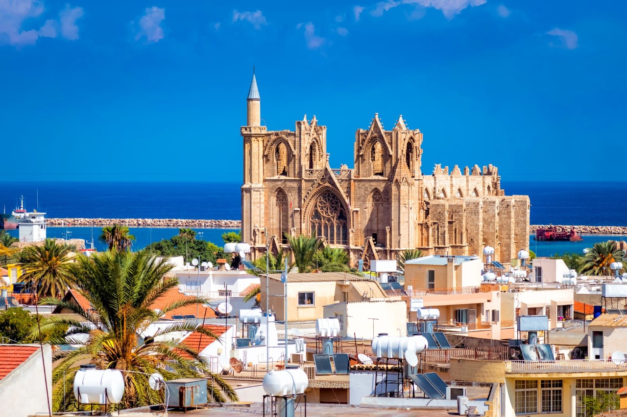 View of a large, sand-coloured building that is a mixture of a Gothic cathedral and a mosque. A minaret rises on the left side of the building and fragments of church towers can be seen in the centre. The building stands in a town and is surrounded by numerous lower houses. There is a palm tree in the left foreground and the sea is visible in the background.