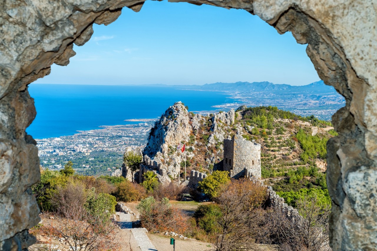 View through masonry to a historic, ruin-like castle complex standing on a hill. A path leads from the foreground towards the castle. Trees and shrubs grow in terraces on the back of the hill. Below the mountain, a sprawling town on a coastline and the sea can be seen.