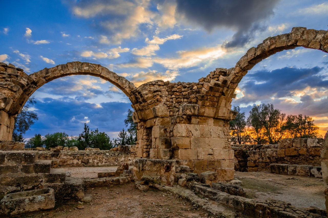 View from the inside of a ruin through two arched gateways, connected by a massive columned ruin, to the sky glowing in the evening, against which a few trees stand out. The ruin is open at the top and provides a view of the sky.