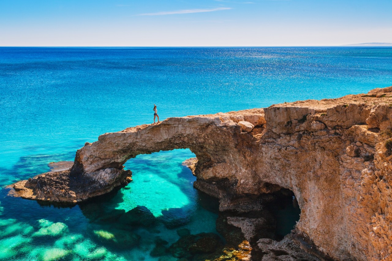 View of a sand-coloured cliff jutting out into the turquoise-blue sea. A section of the cliff leads from the right like a bridge over the sea, on which the silhouette of a woman can be recognised.