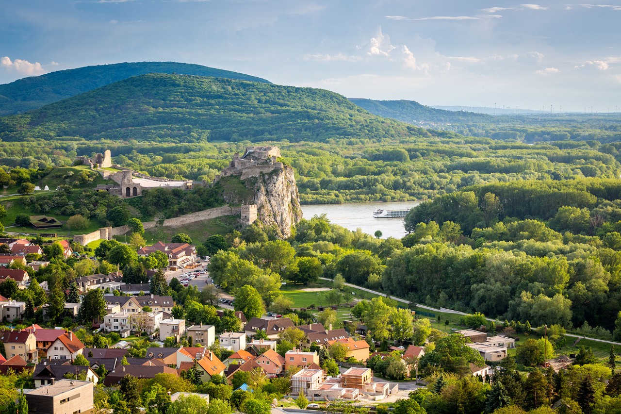 The castle ruins are on the right in the foreground, surrounded by green forests and rivers.