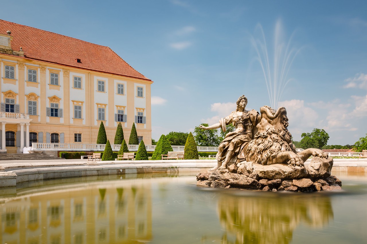 Large fountain with an ornate stone sculpture in a round water basin, in front of a historic castle building with a yellow facade. In the background, symmetrically arranged cone-shaped bushes stand under a blue sky.
