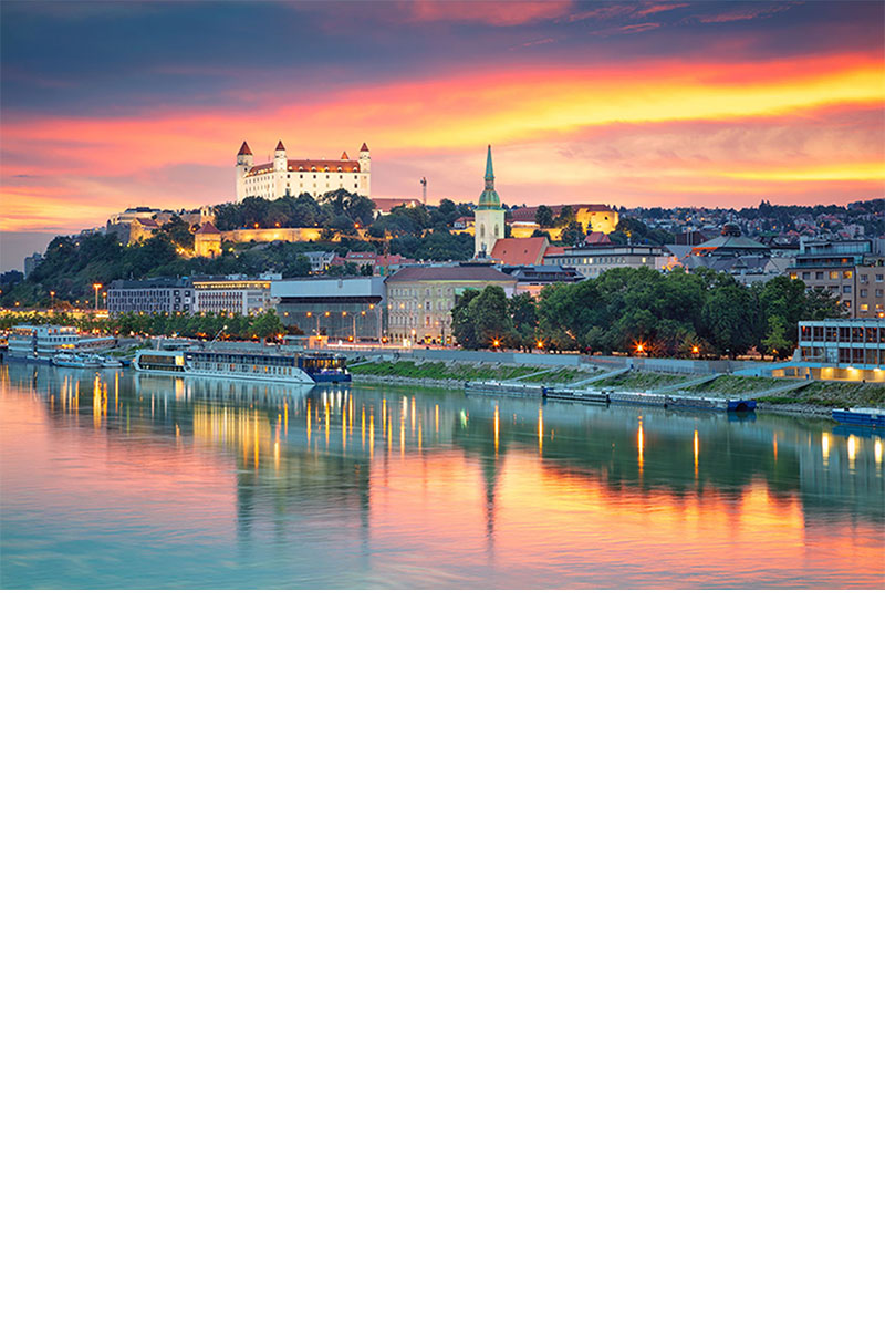Panoramic view of Bratislava at sunset with the illuminated castle on a hill above the city. In the foreground, the city with its church, waterfront promenade, and ships is reflected in the calm waters of the Danube.