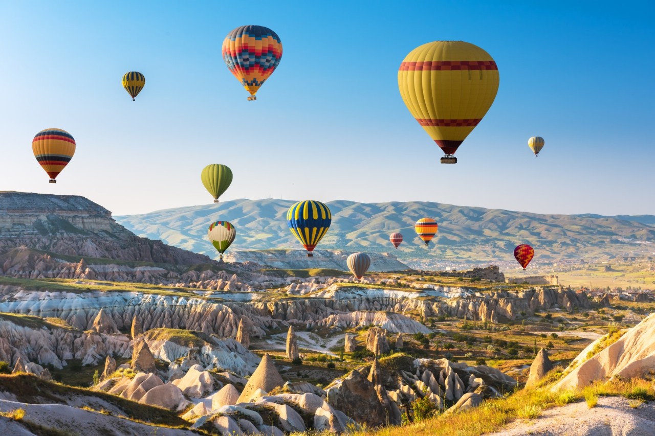 The picture shows numerous colourful hot air balloons floating over a landscape with unusual rock formations and rolling hills. The sky is clear and blue.