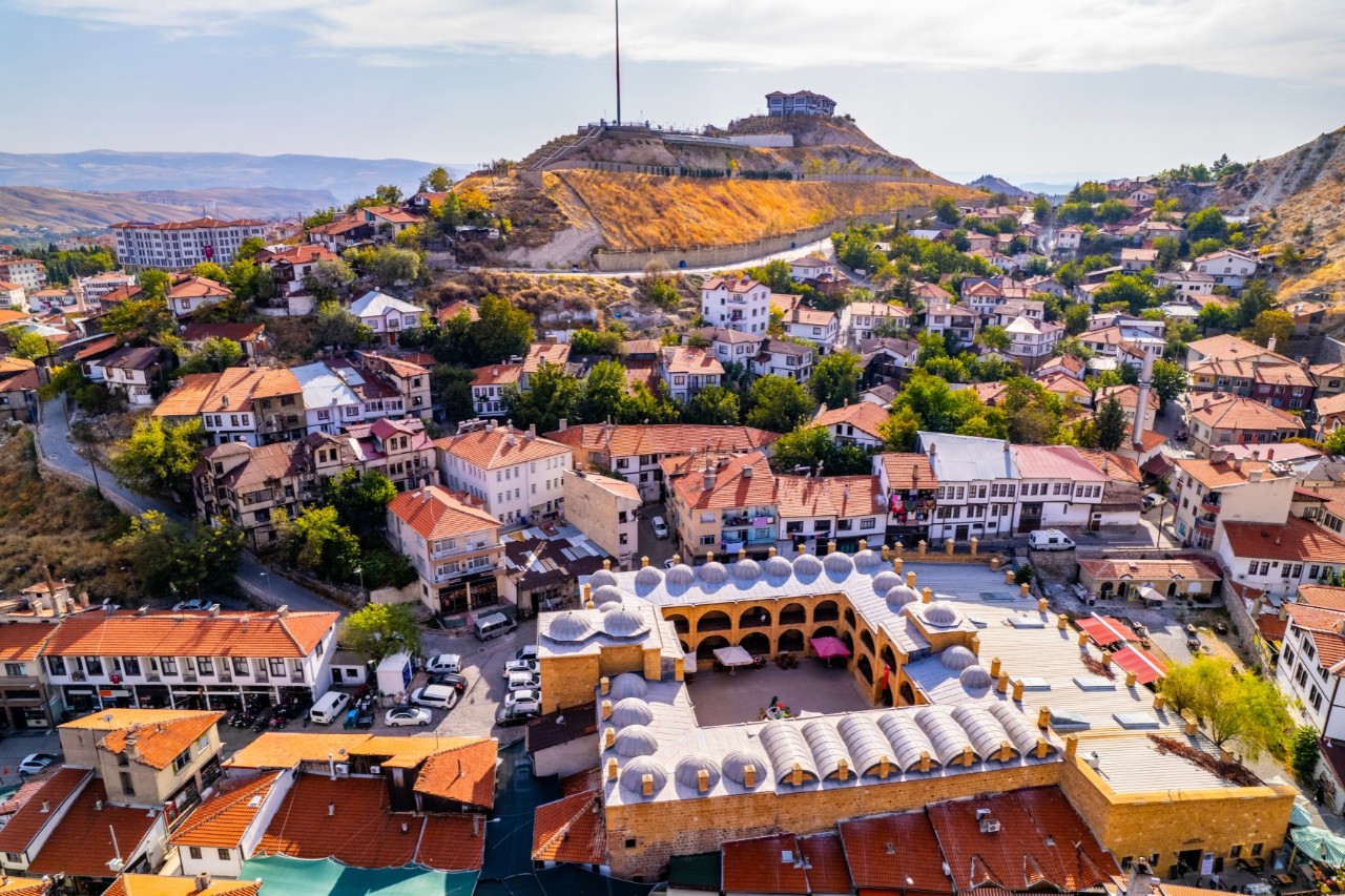 Aerial view of a place with traditional houses and red-tiled roofs. In the foreground, there is a large building with a series of domes. In the background, there is a hill with a fortress. The city is surrounded by green trees and rolling hills.