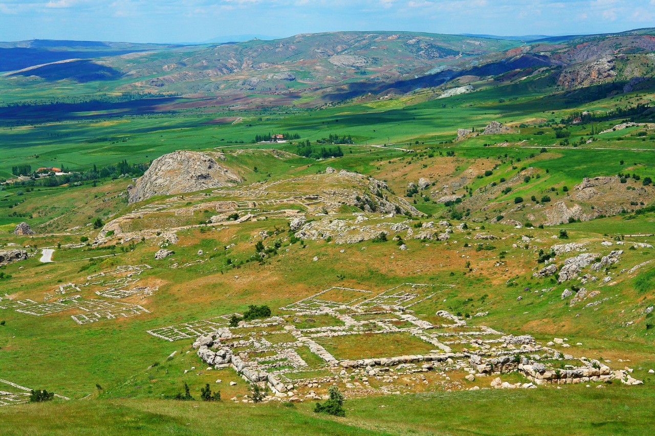 The image shows a wide, green landscape with rolling hills, meadows and stone ruins, which indicate the remains of an ancient city. The wall structures are clearly recognizable and extend over several levels of the hill.