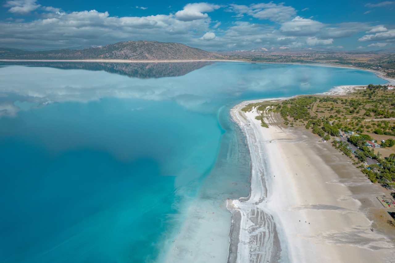 The picture shows a turquoise lake with a white sandy beach. The lake is surrounded by mountains and green vegetation. The water surface reflects the clouds and the sky.