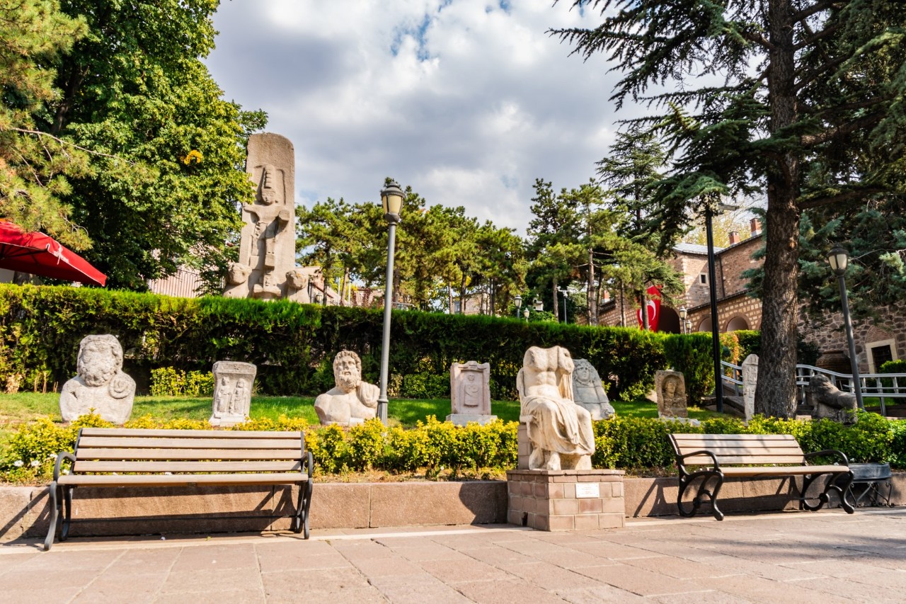 The picture shows a garden decorated with antique statues and busts. There are park benches in the foreground, while trees, old houses and a historic stone wall can be seen in the background.