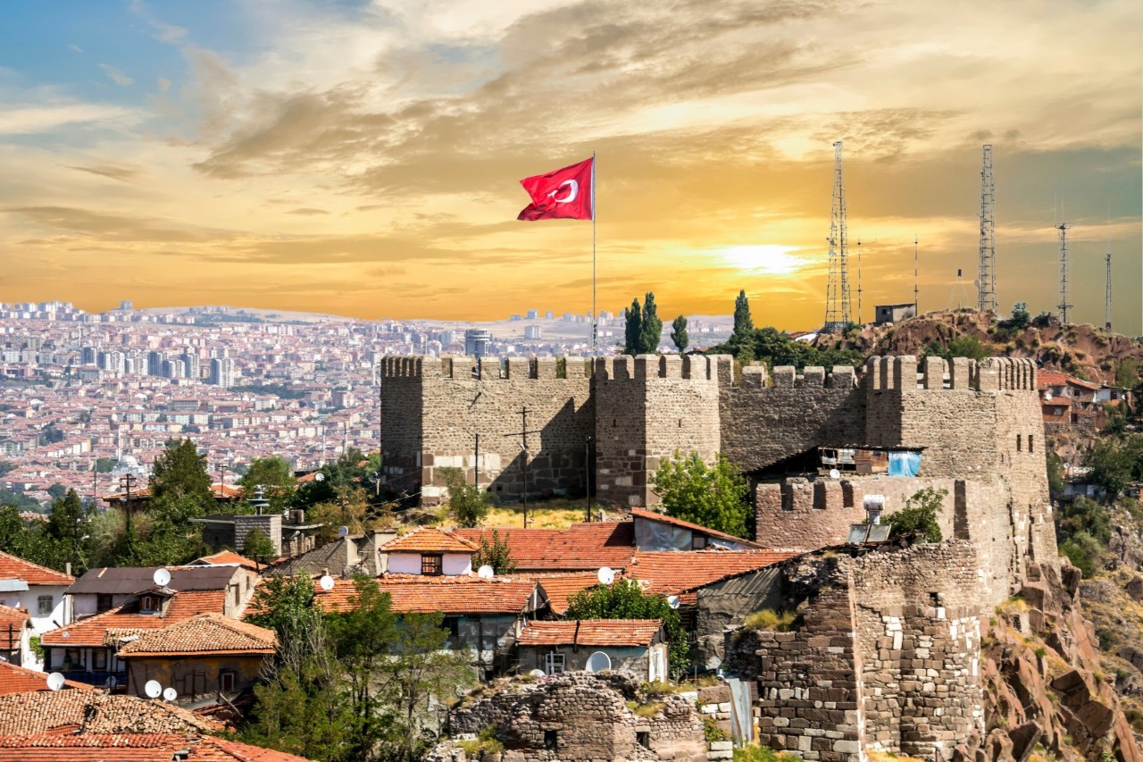 The picture shows a historic fortress with high walls and battlements, with a Turkish flag flying at the top. In the background, a wide urban landscape extends into the distance. The sky glows in shades of yellow and orange at sunset.