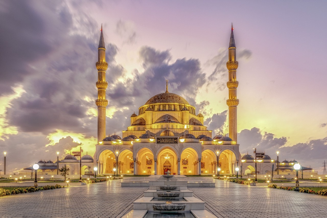 The image shows a large mosque illuminated by warm light. It has a central, impressive dome and four tall minarets. An illuminated square extends in front of the mosque.