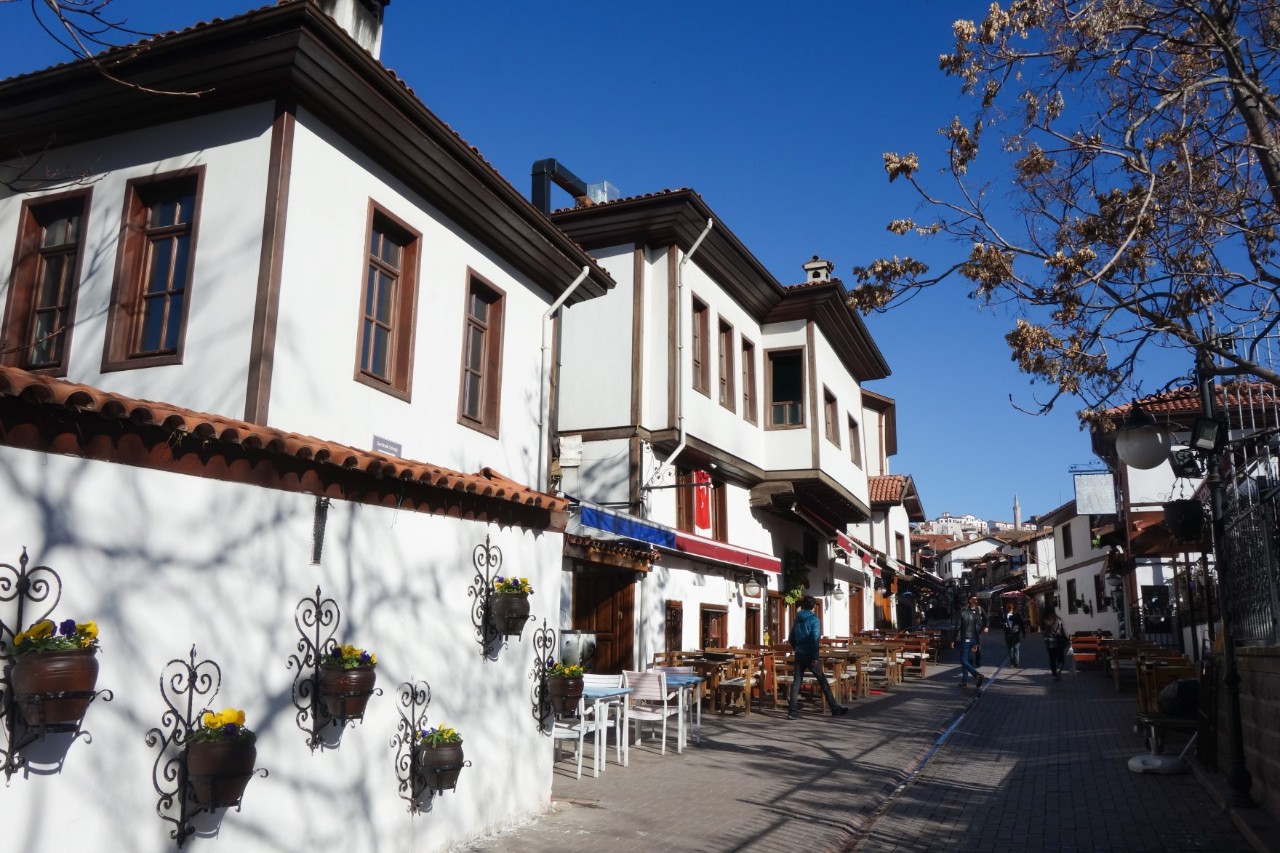 The picture shows white houses along a lane, with wooden elements on the windows and roofs. There are seats along the lane that belong to restaurants down in the buildings. Flower pots are hanging on the walls of the houses.