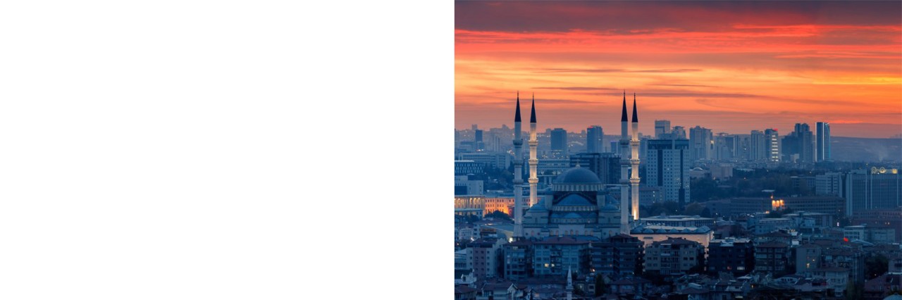 The image shows a cityscape at sunset with an orange-red sky. The focal point is a large mosque with four minarets, while modern high-rise buildings can be seen in the background. 