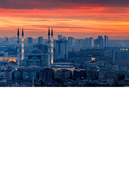 The image shows a cityscape at sunset with an orange-red sky. The focal point is a large mosque with four minarets, while modern high-rise buildings can be seen in the background. 