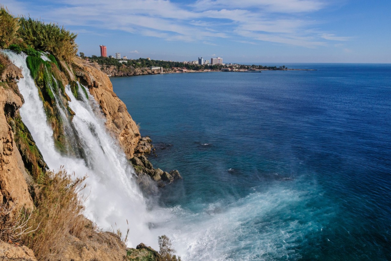 The image shows a large waterfall cascading from a cliff directly into the blue sea. In the background, there is a coastline with scattered buildings and dense vegetation.