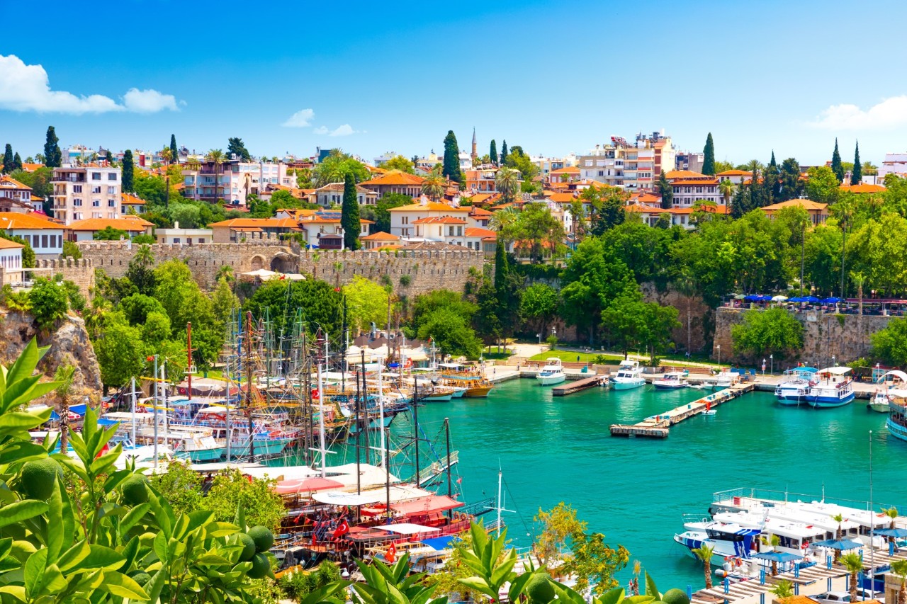 The picture shows an old harbour with boats and sailing ships, surrounded by green trees and the old town with traditional houses and fortress walls in the background.