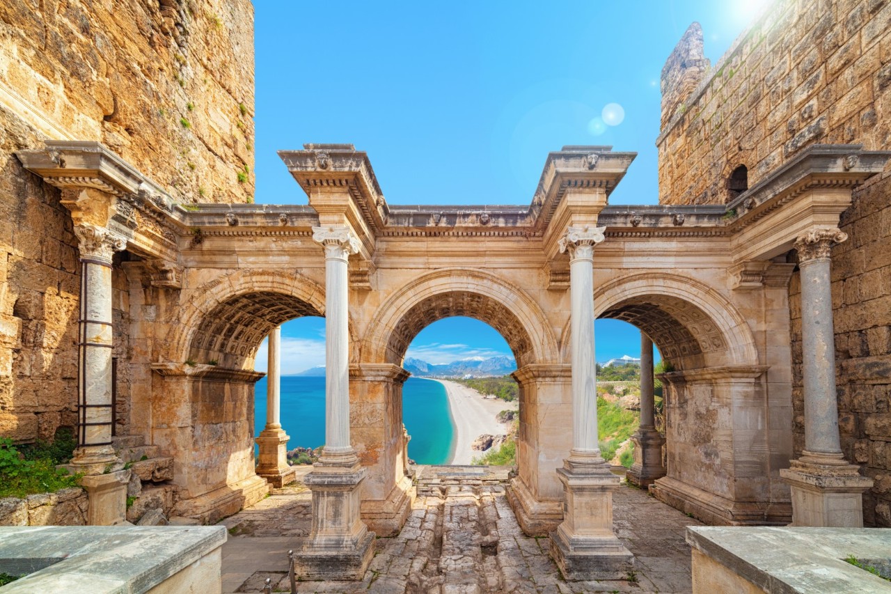 Antique archway with decorated stone columns. Through the arches you can see a coastal landscape with a long beach, turquoise sea and mountains in the background.