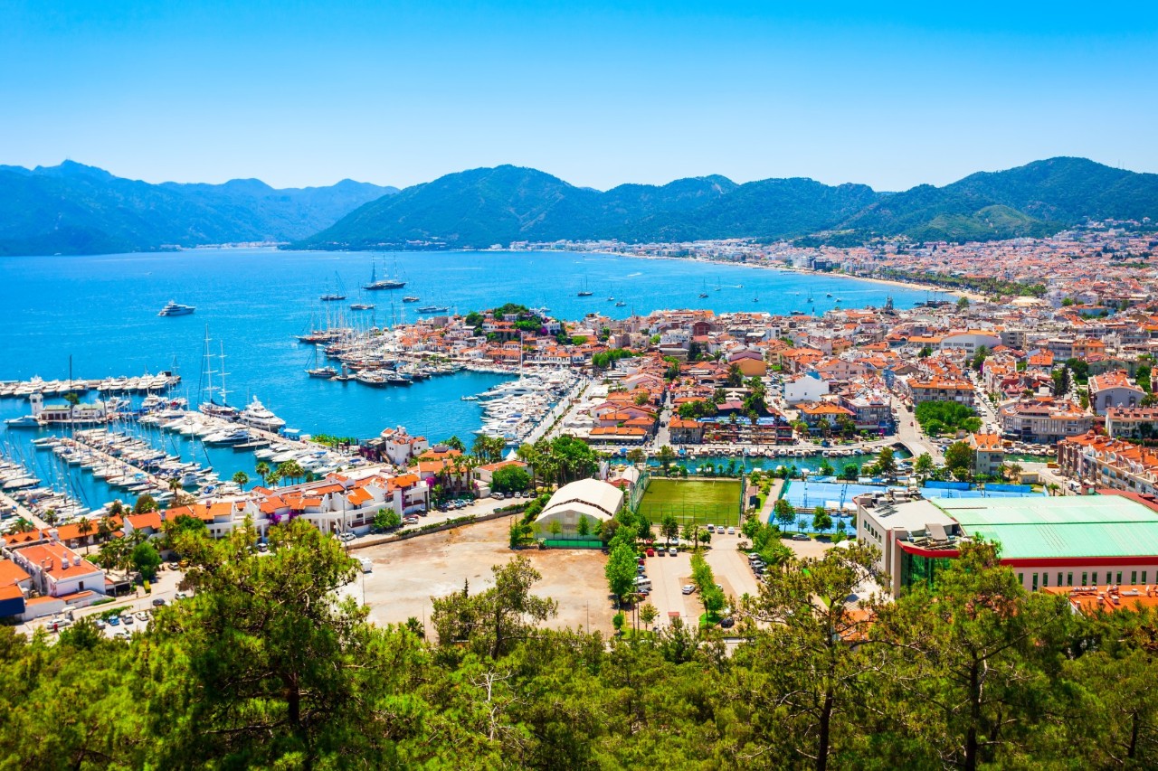 A coastal town with a marina and houses with red roofs directly bordering the turquoise blue sea. Mountains can be seen in the background.
