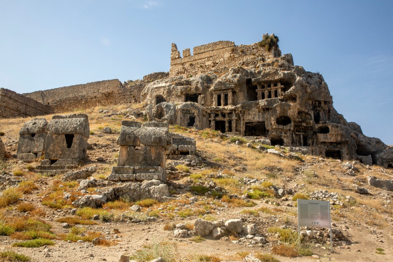 The image shows an ancient ruin on a hillside with several stone sarcophagi and scattered stones in the foreground.