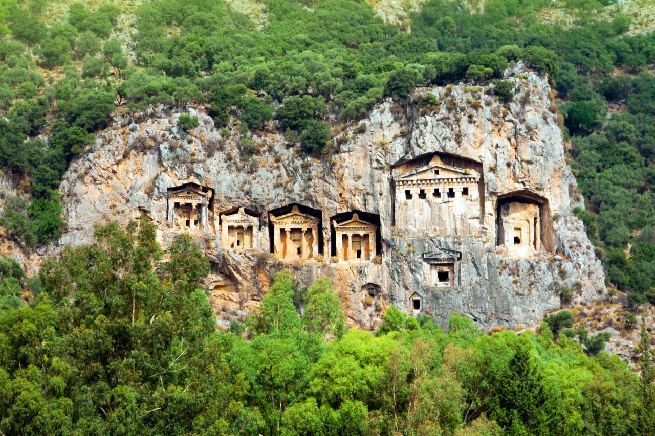 A rock face with several rectangular burial chambers carved into the stone. Their facades are column-like. Trees and dense vegetation enclose the rock.