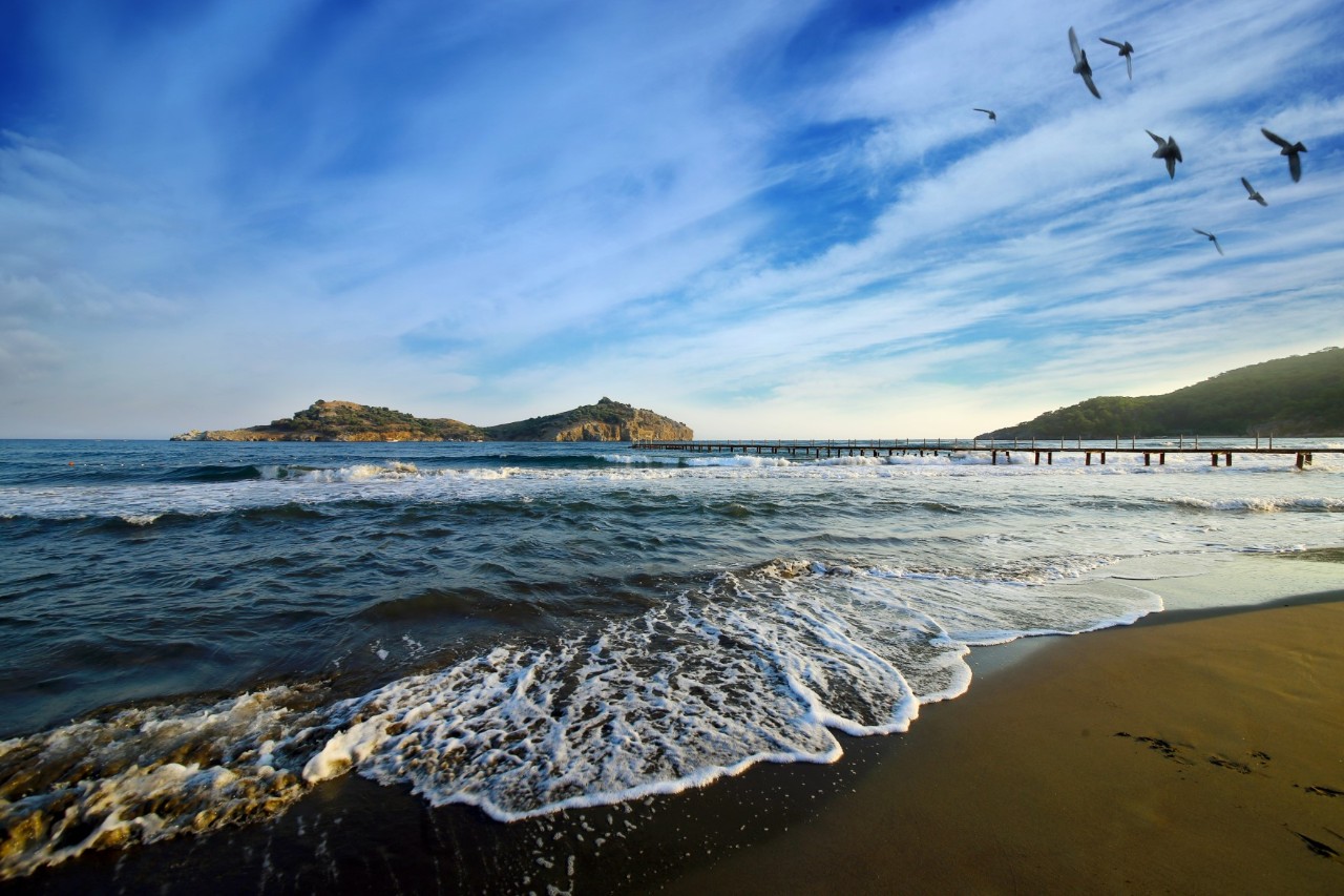 An ochre-coloured sandy beach by the dark blue sea with gentle waves and seagulls in the sky. In the background, a small wooden bridge and two wooded island hills can be seen.