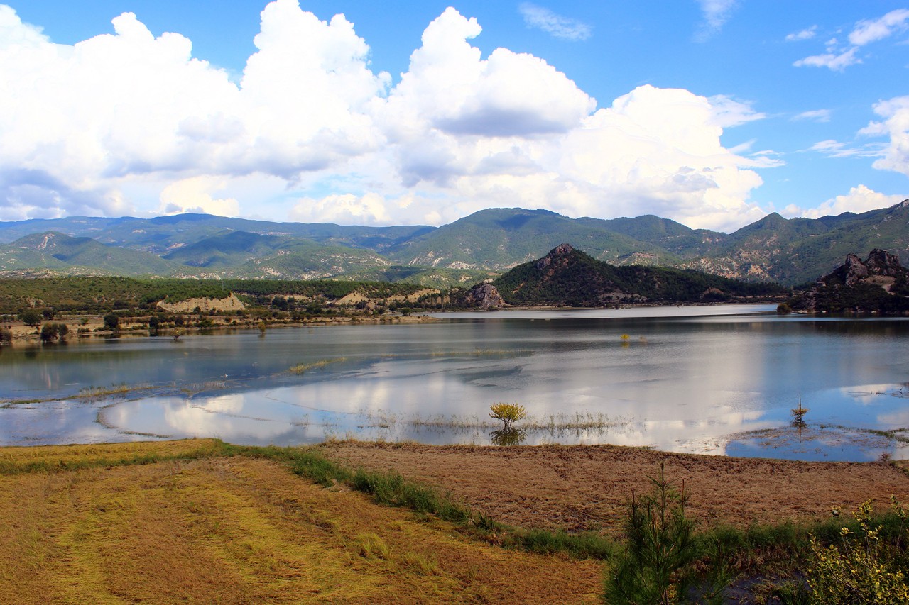 Quiet lake landscape with flat shore in the foreground and green mountains in the background. The sky is partly cloudy.