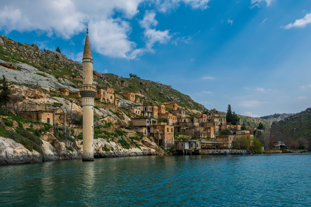 Houses painted in shades of yellow on a mountainside on the banks of a wide river. A single minaret rises out of the water.