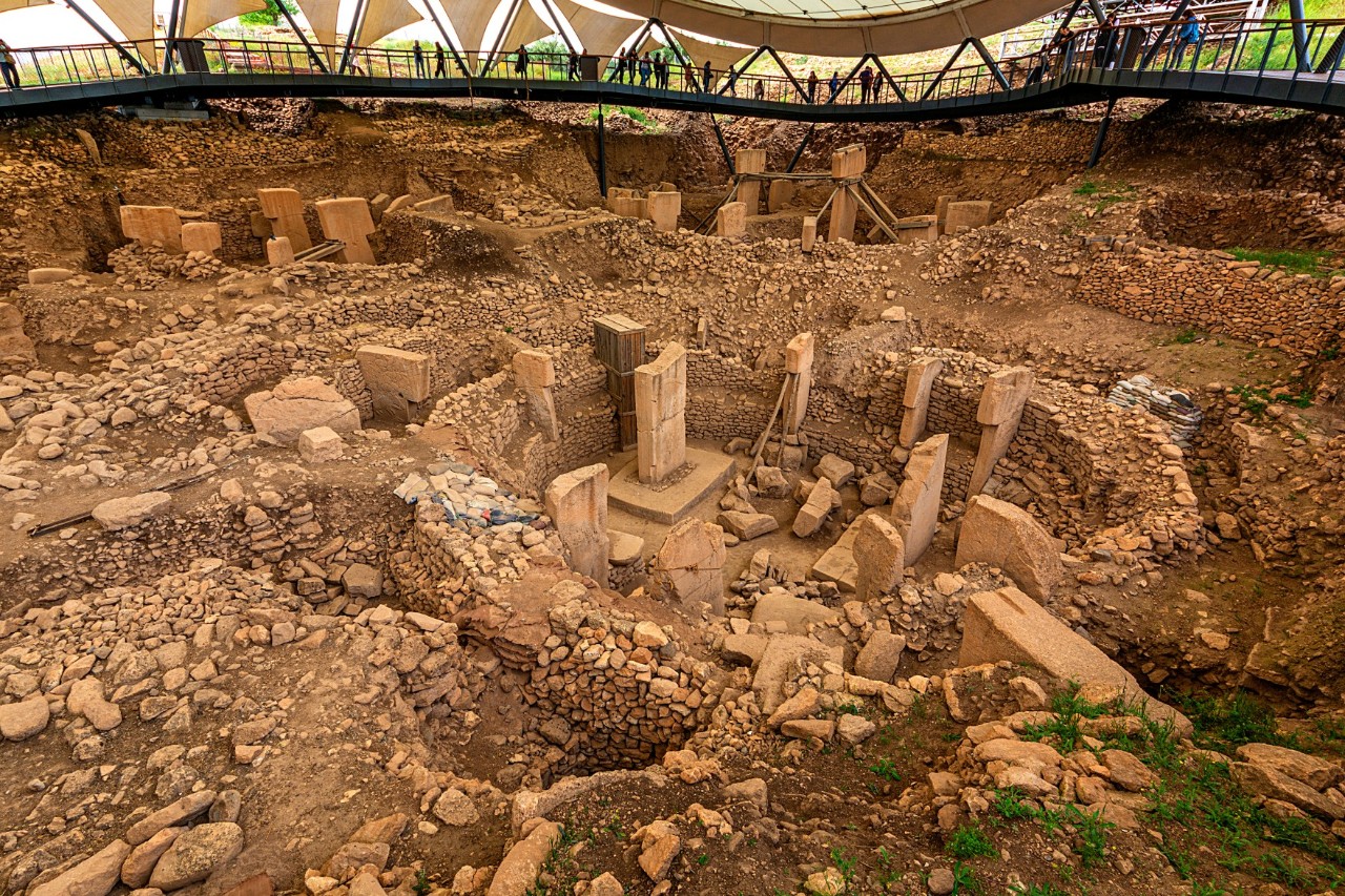 An archaeological excavation site with large stones arranged in a circle, covered by a structure with a visitor platform.