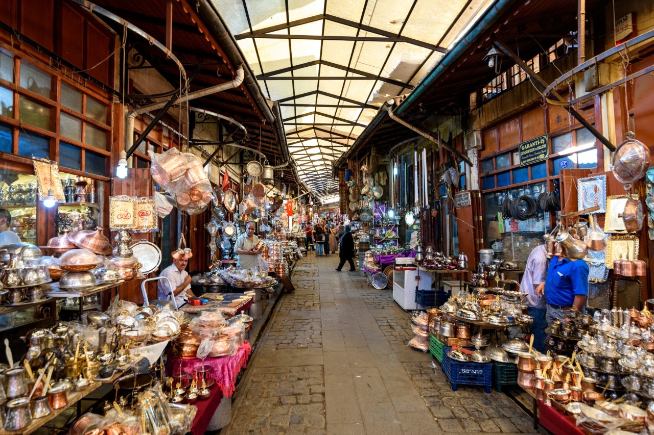 Covered bazaar with cobblestones and numerous stalls selling copper and silverware. People stroll through the alley.