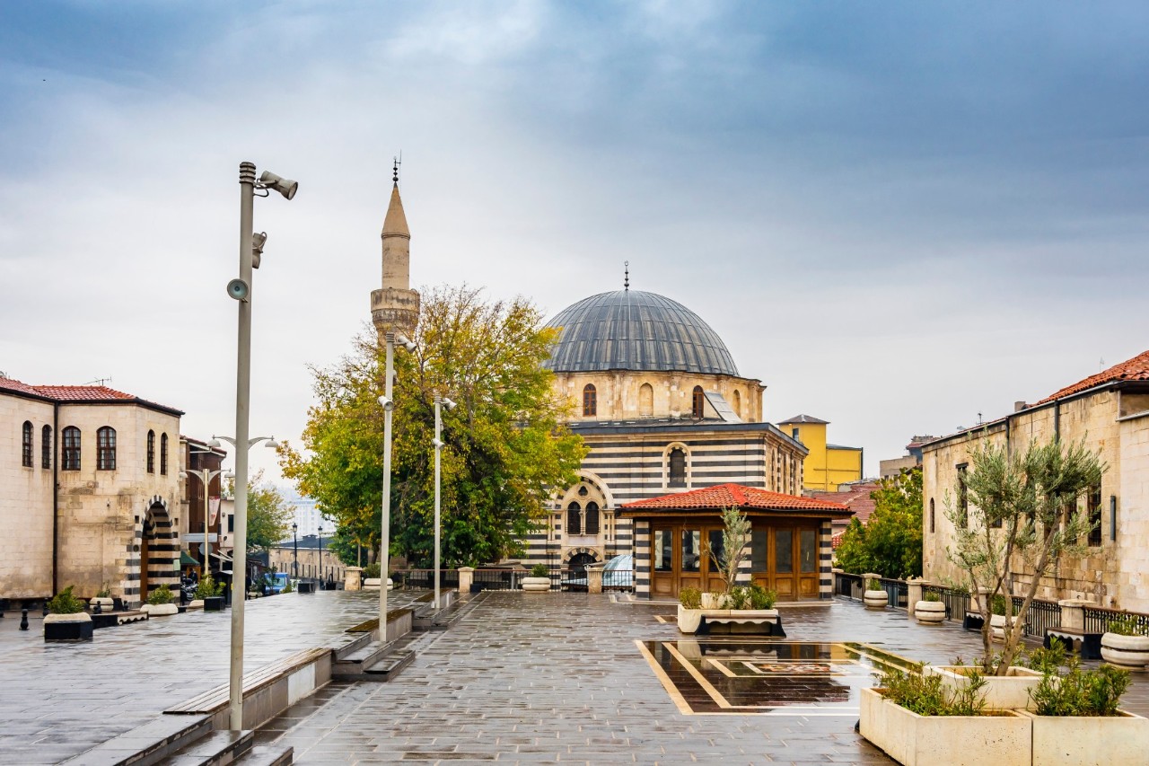 View of a historic mosque with a striped façade, large dome and minaret, surrounded by a paved square with trees and benches.