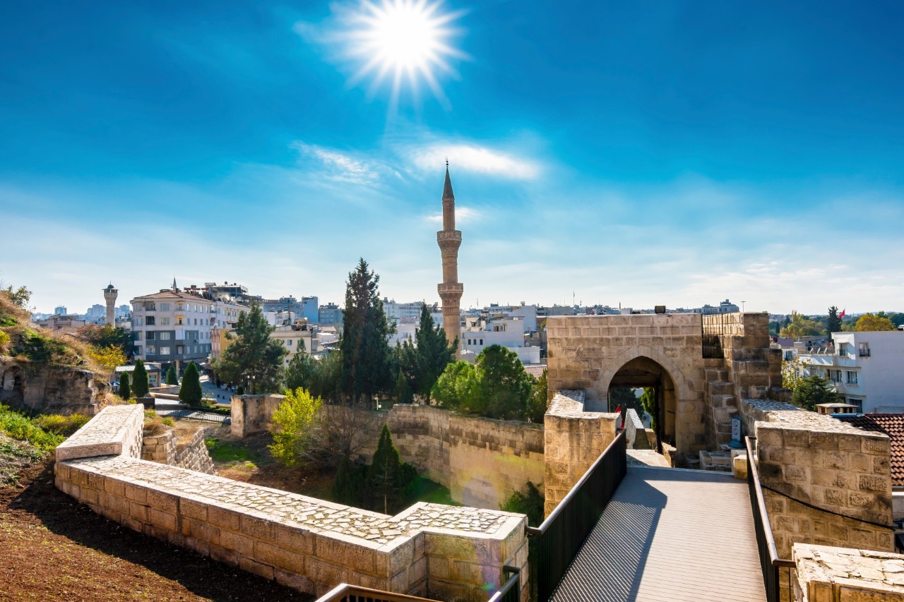 View of a city with a stone castle in the foreground, a minaret in the centre, bright houses and a clear blue sky.