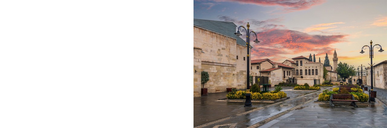 Paved square with Ottoman architecture, yellow flower beds, street lamps and a mosque with a minaret in the background. The sky shows scattered clouds in shades of red and yellow.