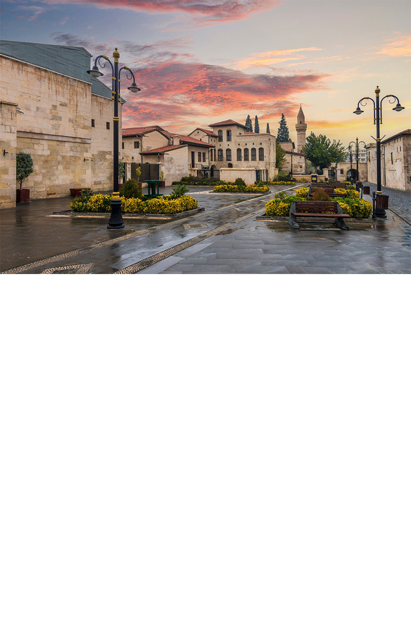 Paved square with Ottoman architecture, yellow flower beds, street lamps and a mosque with a minaret in the background. The sky shows scattered clouds in shades of red and yellow.