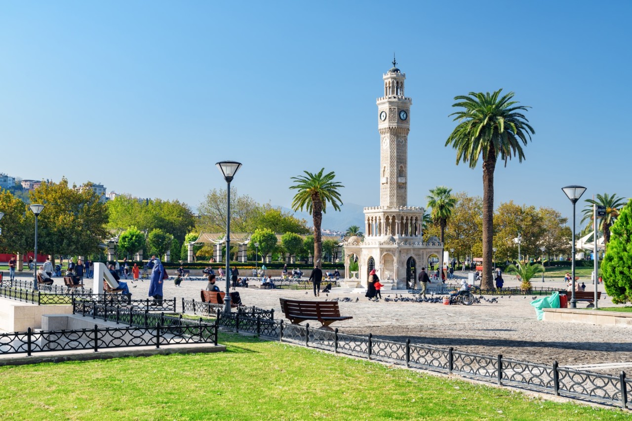 View across a green lawn surrounded by a low metal fence to a square with a bright, tall clock tower in the centre. The square is lined with palm trees and benches, with several people strolling around.