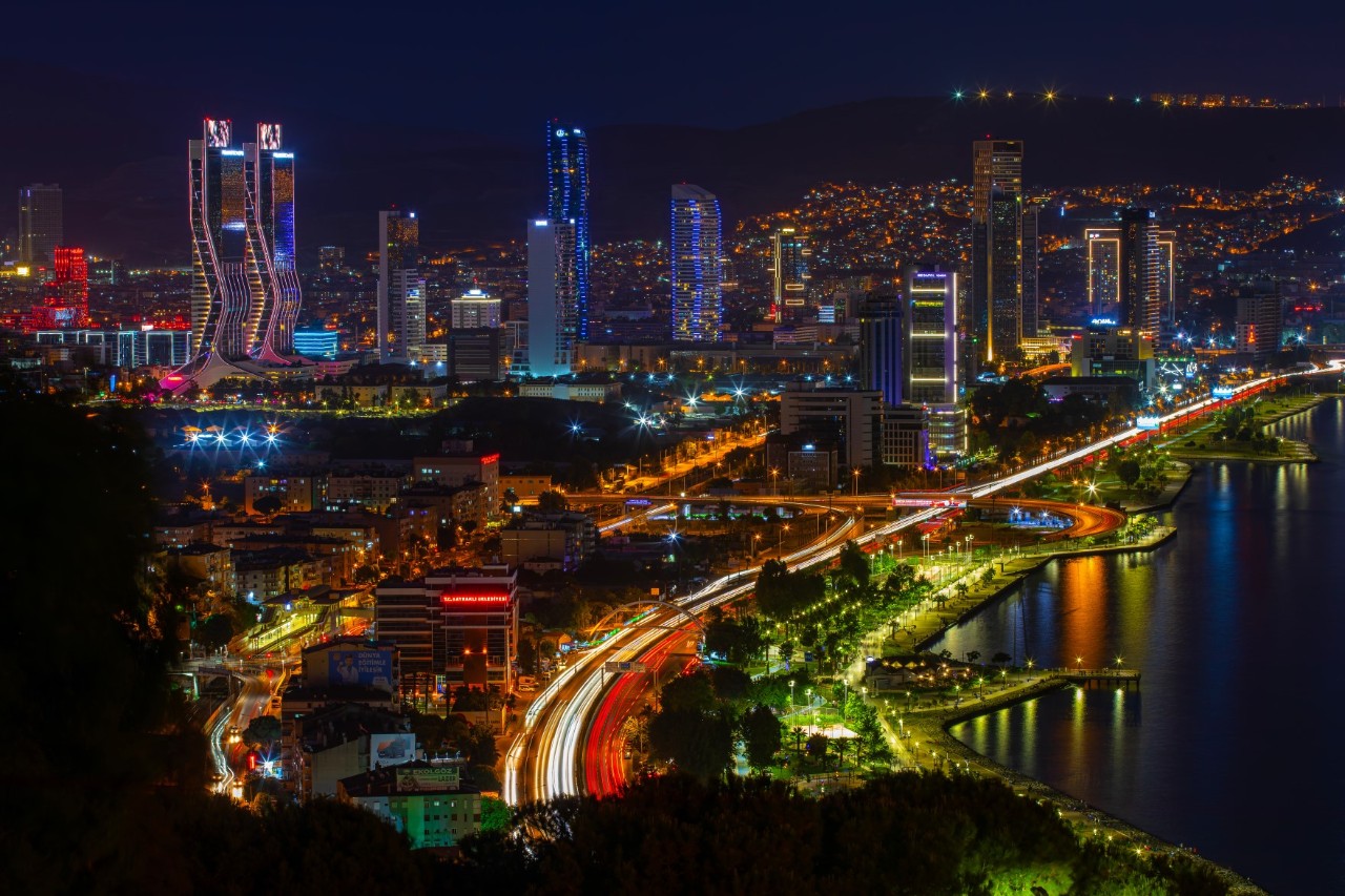 Night shot of a colourfully illuminated city from a bird's eye view. A floodlit street can be seen on the right-hand side of the picture, while the skyline with numerous illuminated skyscrapers can be seen in the background.