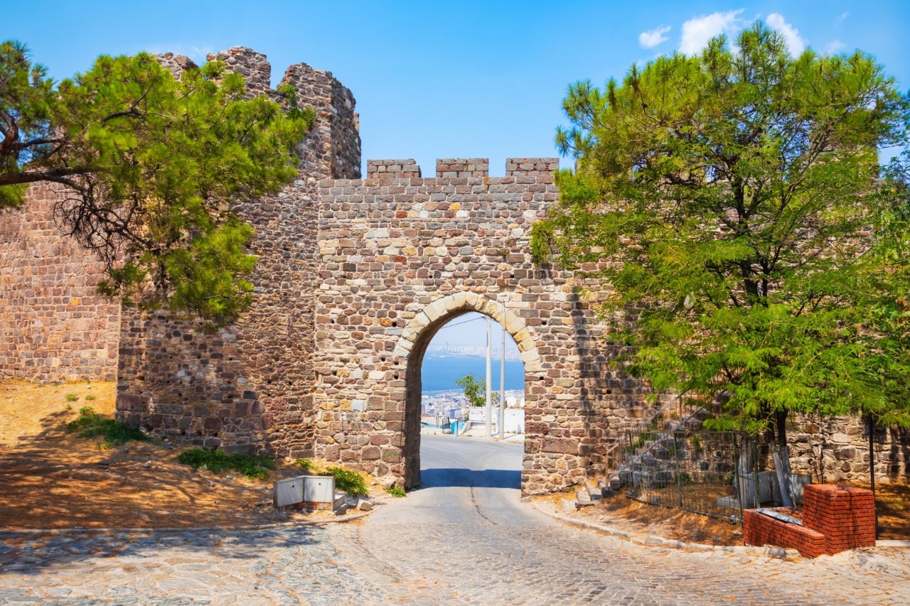 View of a castle wall amid battlements and a defence tower next to an open brick city gate and a cobbled street. Parts of a town and the sea can be seen through the open gate. A flight of steps leads up to the right in front of the gate. There are trees in the foreground. The sky is blue.