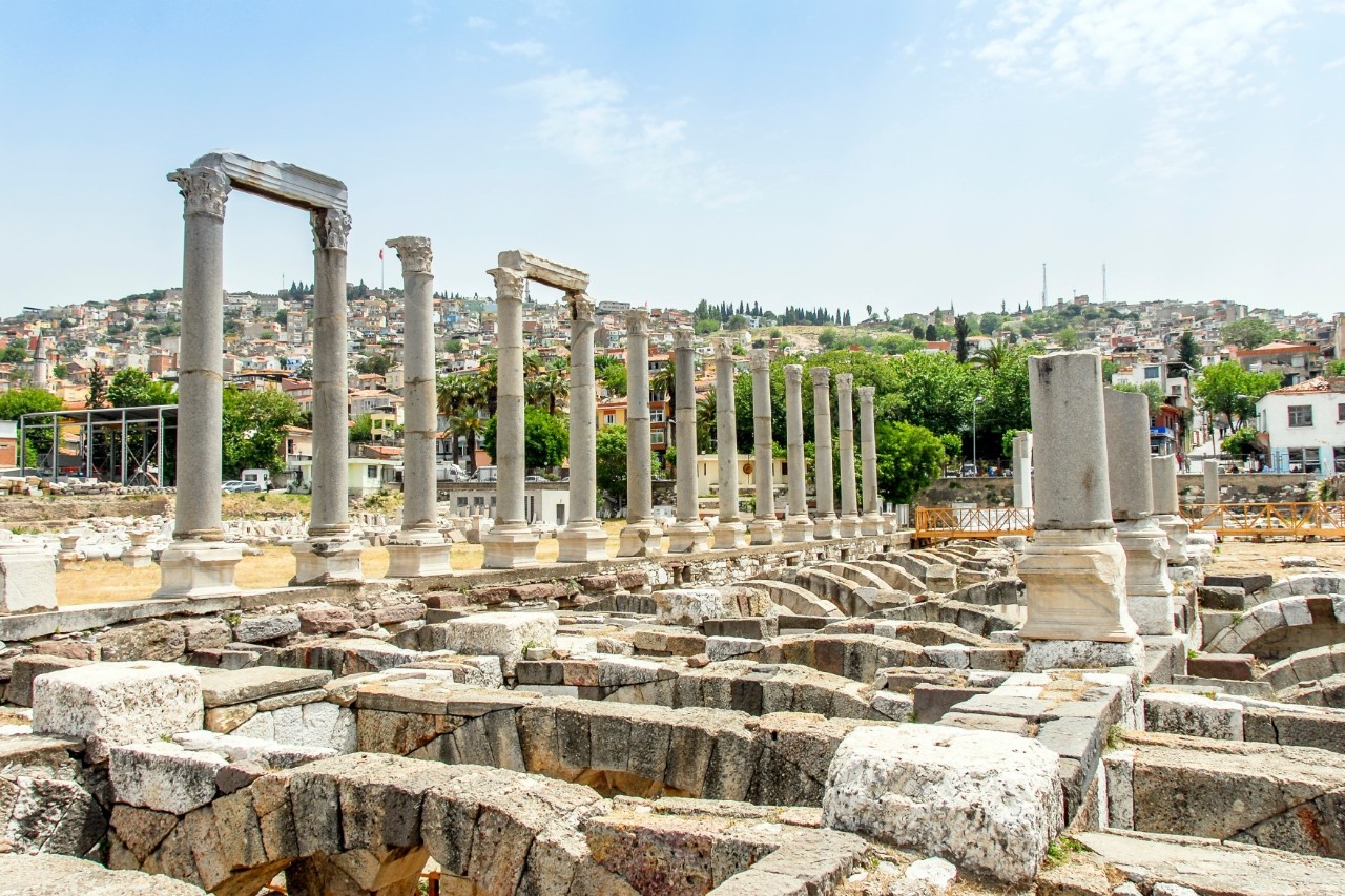 View of an archaeological site comprising several remains of walls and light grey columns. A city on a hill can be seen in the background.