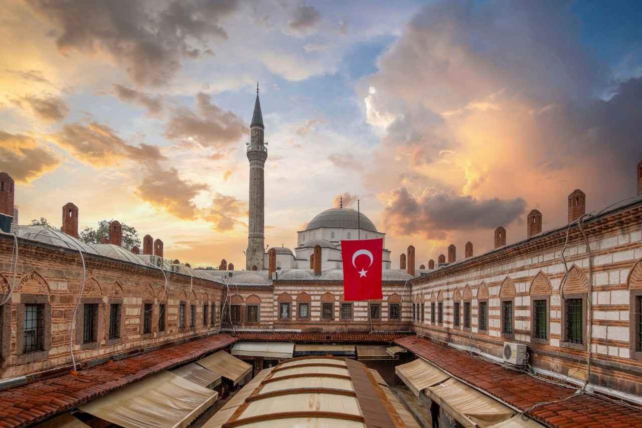 View of an inner courtyard surrounded by a reddish-brown and white wall with windows. The dome and a high minaret of the mosque are visible in the background, with a Turkish flag hanging in front of it. The inner courtyard is covered by an arched roof and awnings on the sides. In the background, the slightly cloudy sky glows in the sunset.