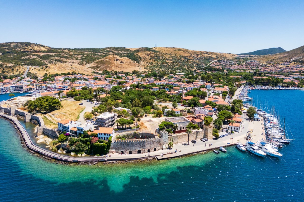 Aerial view of an urbanised headland surrounded by the blue sea. A fortress wall and a harbour with several small boats can be seen in the foreground. The houses are light-coloured with red roofs, with many trees visible in between. The town continues behind the headland and merges into hilly mountainous terrain. The sky is blue.