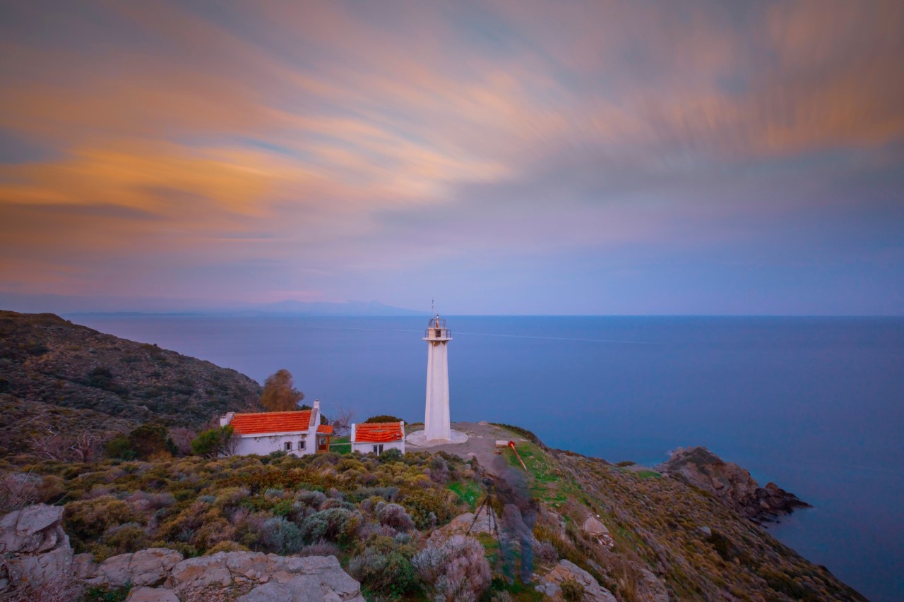 View of a cliff by the sea at sunset. There is a white lighthouse on the cliff. Two white houses with bright red roofs can be seen to the left of the lighthouse. Behind the cliff. you can see the sea.