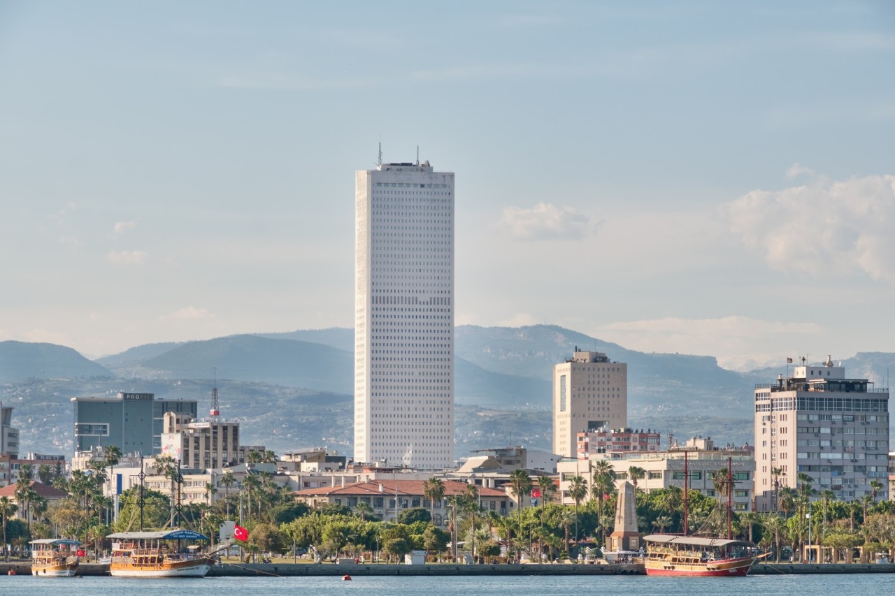 View from the opposite bank of a city with a striking white skyscraper in the center, surrounded by smaller buildings, palm trees, and boats along the coast. Mountains rise in the background.