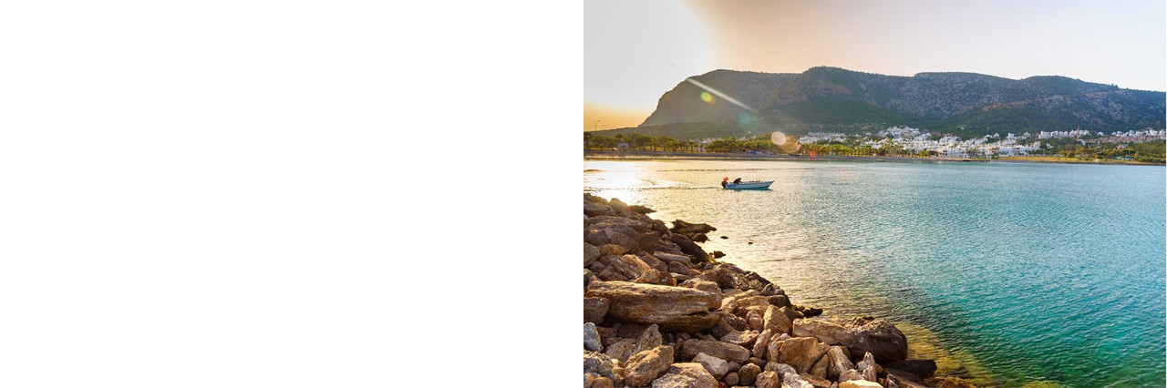 A small boat sails across the calm, turquoise sea at sunset in front of a coastal town with white houses, with wooded mountains in the background.