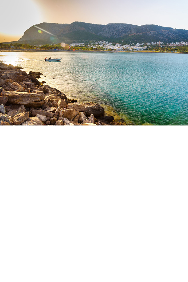 A small boat sails across the calm, turquoise sea at sunset in front of a coastal town with white houses, with wooded mountains in the background.