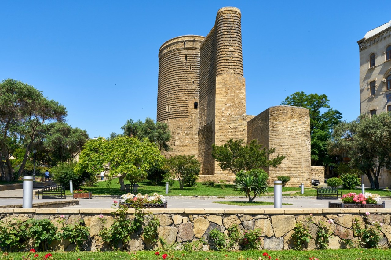 In the centre of the picture, there is a massive, round tower made of sand-coloured stone with a protruding, semicircular part of the building and another extension to the right of it on a square. The building is surrounded by trees. On the right side, a multistorey building protrudes from the picture.