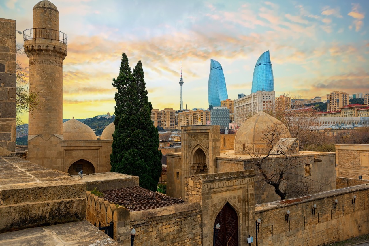 Sand-coloured, massive walls, several domes and a round tower of a palatial complex rise up in the foreground. In the courtyard stand two entwined conifers. Multiple arched doorways and lancet windows break up the walls. In the background are residential buildings, as well as two flame-shaped high-rises and a television tower.